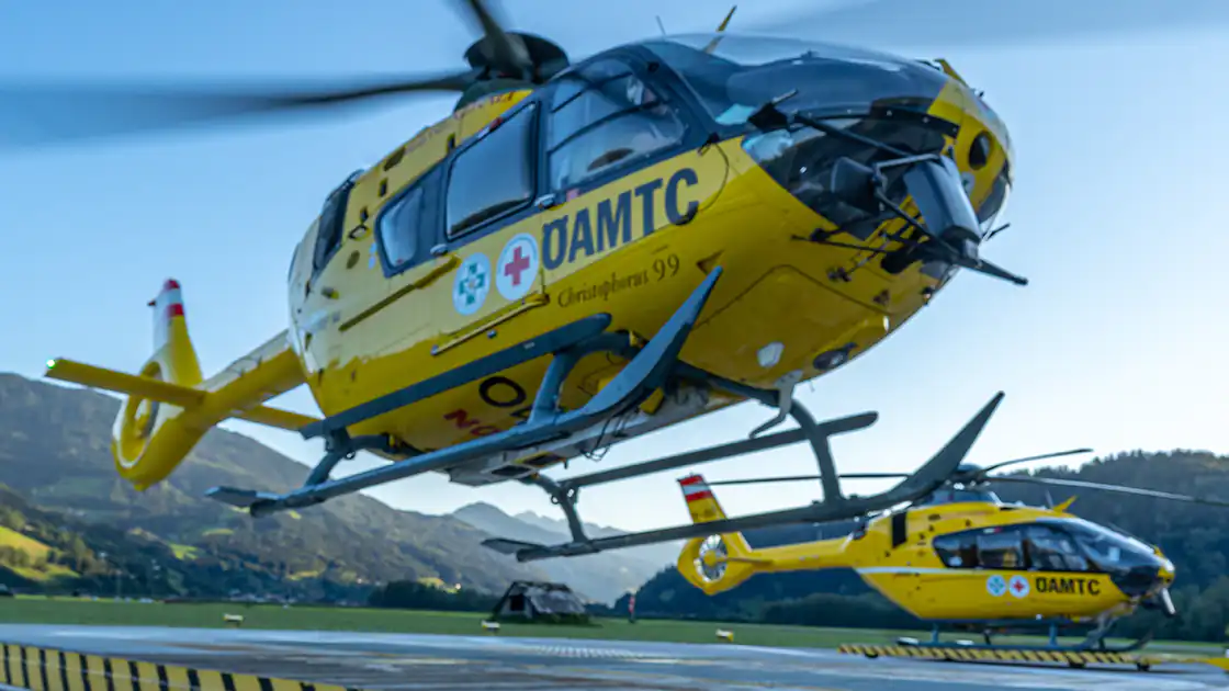 Zwei Rettungshubschrauber auf einem Hubschrauber-Landeplatz. Ein Rettungshubschrauber ist schon gestartet, der zweite steht noch auf dem Boden. Im Hintergrund sind Berge zu sehen.
