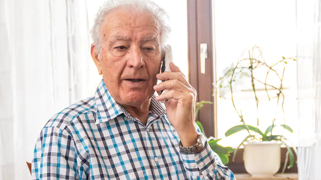 Ein Symbolbild zeigt einen älteren Mann,der mit einem drahtlosen Telefon telefoniert. Er sitzt dabei vor einem Fenster. Zu sehen ist eine Zimmerpflanze neben ihm auf dem Fensterbrett. Der ältere Mann trägt ein kariertes Hemd und eine glitzernde Armbanduhr.