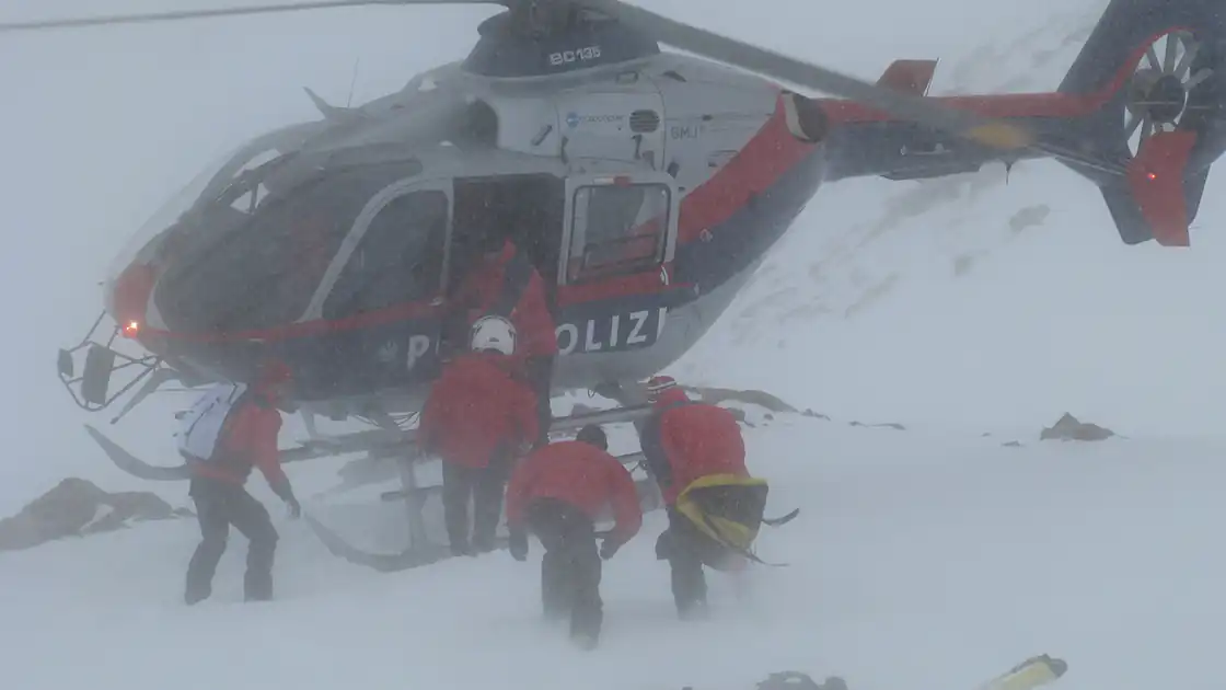 Ein Alpineinsatz der Polizei ist abgebildet. In einer schneeverwehten Umgebung mit Nebel steht ein Poilzeihubschrauber in den Bergen. Rundherum ist Schnee, mehrere Polizisten im Einsatz stehen an der geöffneten Tür des Helikopters.