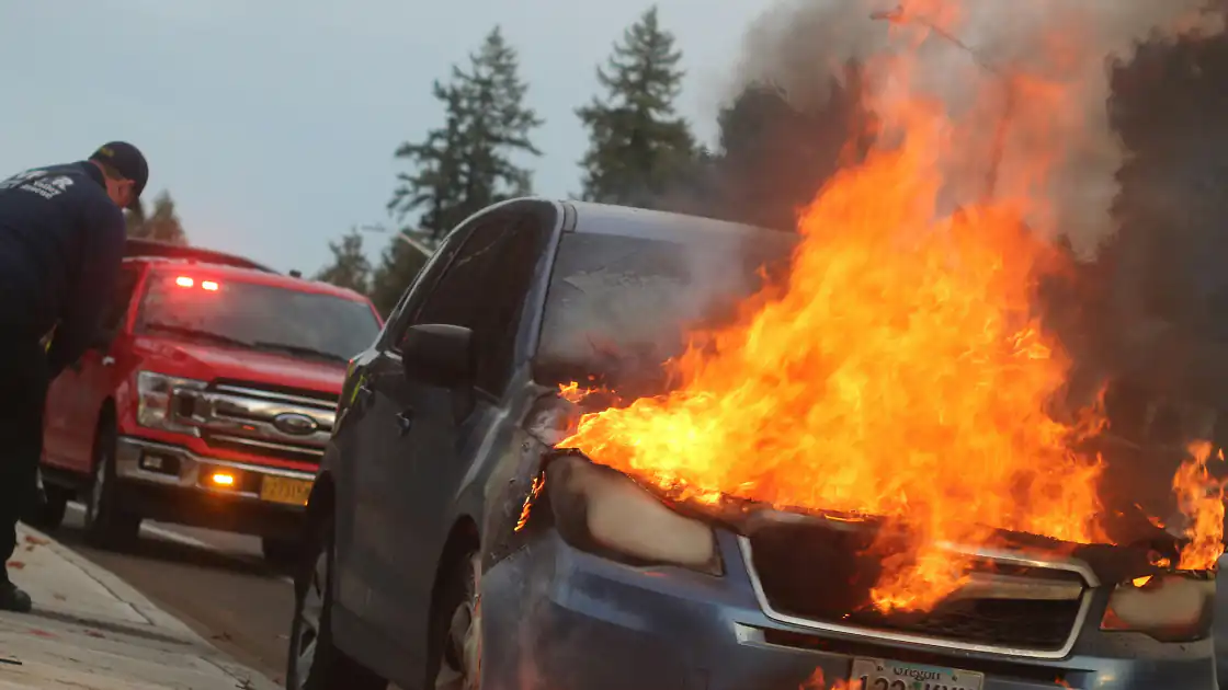 Ein Auto / PKW im Vollbrand ist in diesem Symbolphoto zu sehen. Dahinter steht bereits ein Einsatzfahrzeug. Der Wagen brennt lichterloh aus dem Motorraum.