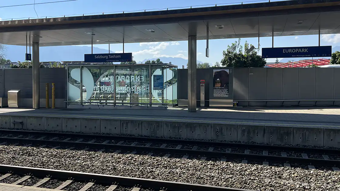 Am Bild sind zwei Gleise und dann der zweite Teil des Bahnhofsgebäudes in Salzburg Taxham beim Europark zu sehen. Europark ist ein großes Einkaufszentrum in der Stadt Salzburg.