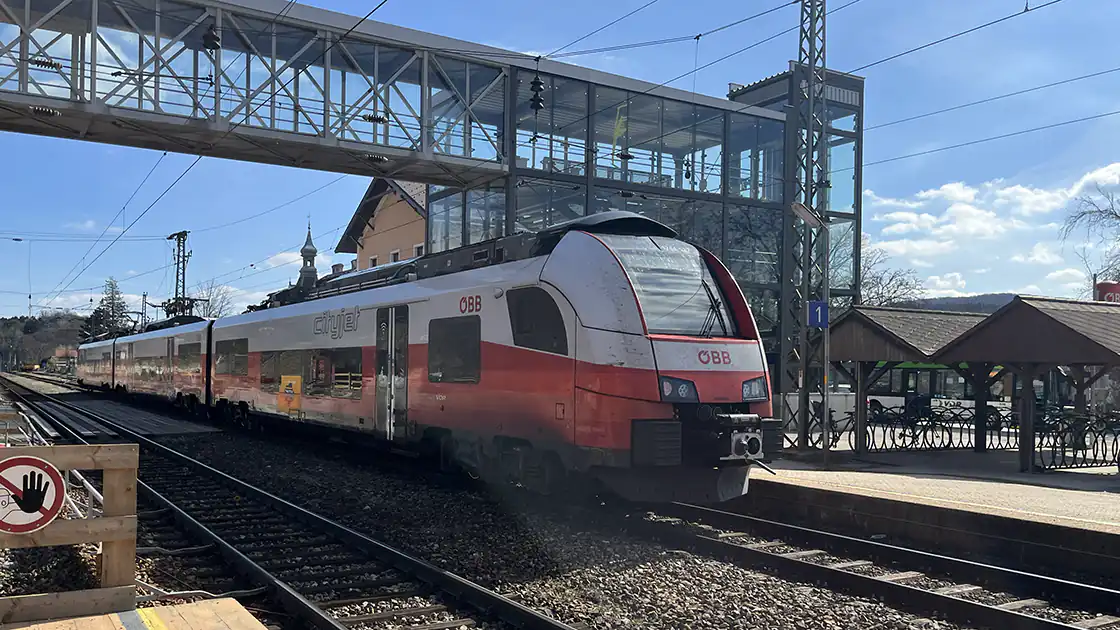 Der Bahnhof Tullnerbach-Pressbaum in Niederösterreich ist hier im Bild zu sehen. Die beiden Gleise sind von einer Fußgängerüberführung, die mit einer Glas-Röhre gemacht ist, umrahmt. Unter der Unterführung fährt gerade eine Eisenbahn auf dem Gleis rechts im Bild durch. Das linke Gleis is frei. Neben dem Lift und dem Aufgang zur Überführung ist ein offener aber überdachter Bereich als Fahrradabstellplatz zu sehen.
