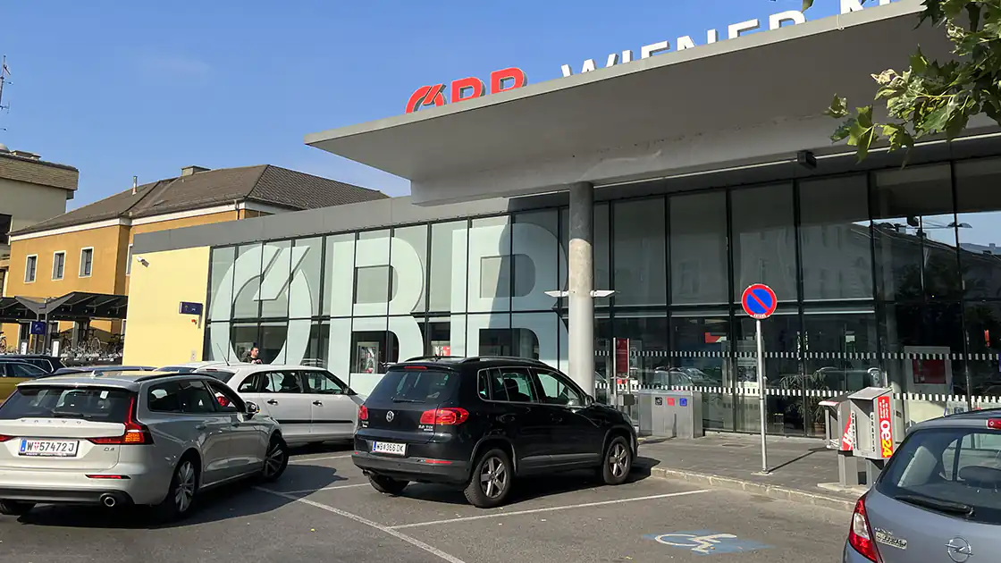 Die Front-Ansicht des Bahnhof Wiener Neustadt, vor dem isch direkt ein kleiner Parkplatz befindet. Hier im Gebäude geht es hinein in den Bahnhof, wo sich auch ein kleiner Markt, Bäckerei, Trafik und ähnliche Einrichtungen befinden. Wenn man dann über die Rolltreppe hinunterfährt, kann man zu den weiteren Bahnsteigen gelangen.