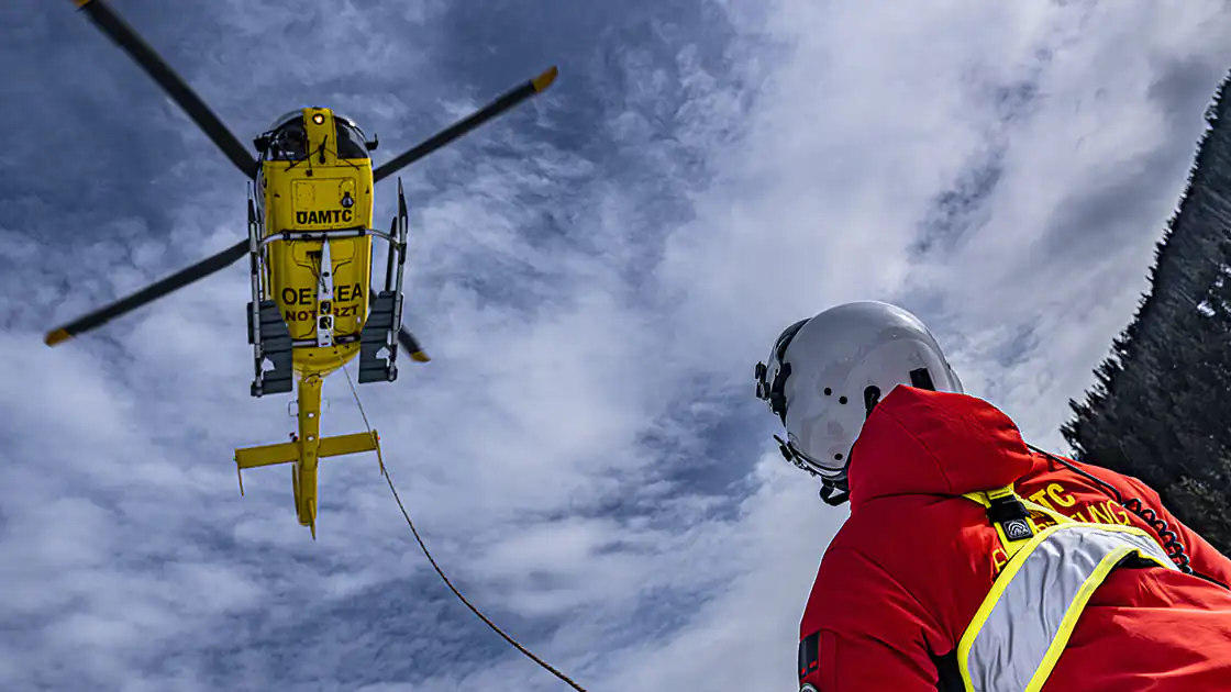 Ein Rettungshubschrauber ist von unten zu sehen, er hat ein Seil heruntergelassen. Am anderen Bildrand steht eine Person, die zum Rettungsteam gehört und am Boden für die Fixierung des Unfallopfers zuständig ist.