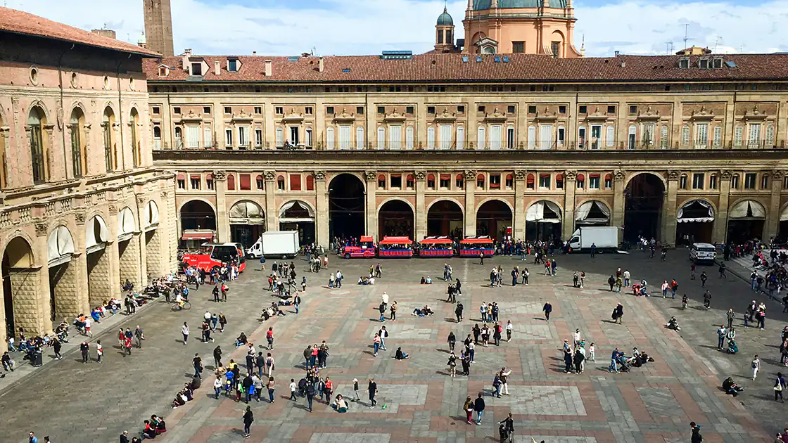 Ein großer Platz in der italienischen Stadt Bologna ist im Bild mit vielen kleinen Menschen zu sehen. Rund um den Platz sind alte Gebäude mit Säulen und Arkaden zu sehen.