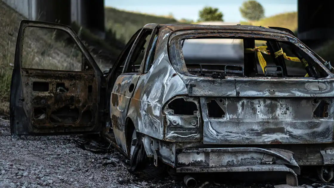 Wenn ein Auto völlig ausgebrannt ist, dann sieht das etwa so aus. Im Bild ist ein weißes völlig ausgebranntes Auto-Wrack mit einer geöffneten Fahrertür auf der linken Seite zu sehen.