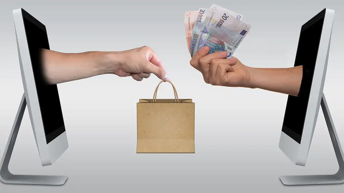 E-commerce-Symbolbild: Zwei Computermonitore stehen auf einer weißen Fläche einander gegenüber. Aus einem Monitor ragt eine Hand mit einer braunen Papiertasche heraus, aus dem anderen Monitor kommt eine Hand mit einem Bündel Bargeld entgegen.