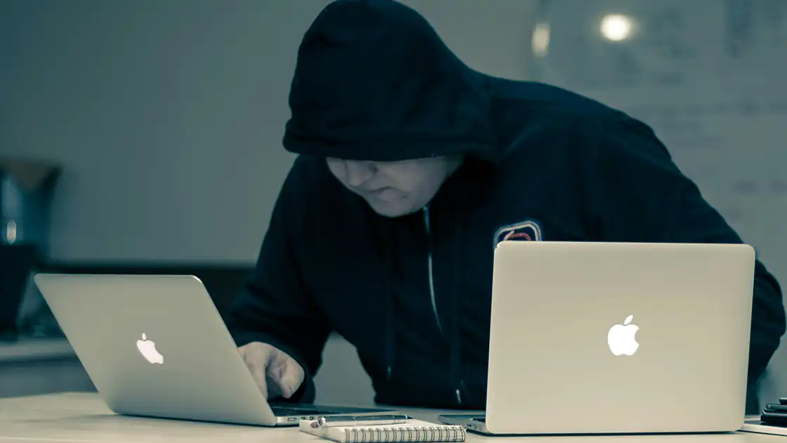 Finstere Gestalten in der Online-Welt machen sich immer wieder an Leute heran und überzeugen diese von Dingen, die man normalerweise nicht machen sollte. Das Bild stellt einen typischen Hacker dar, der mit einem schwarzen Kapuzen-Shirt vor zwei Laptops der Marke Apple sitzt.