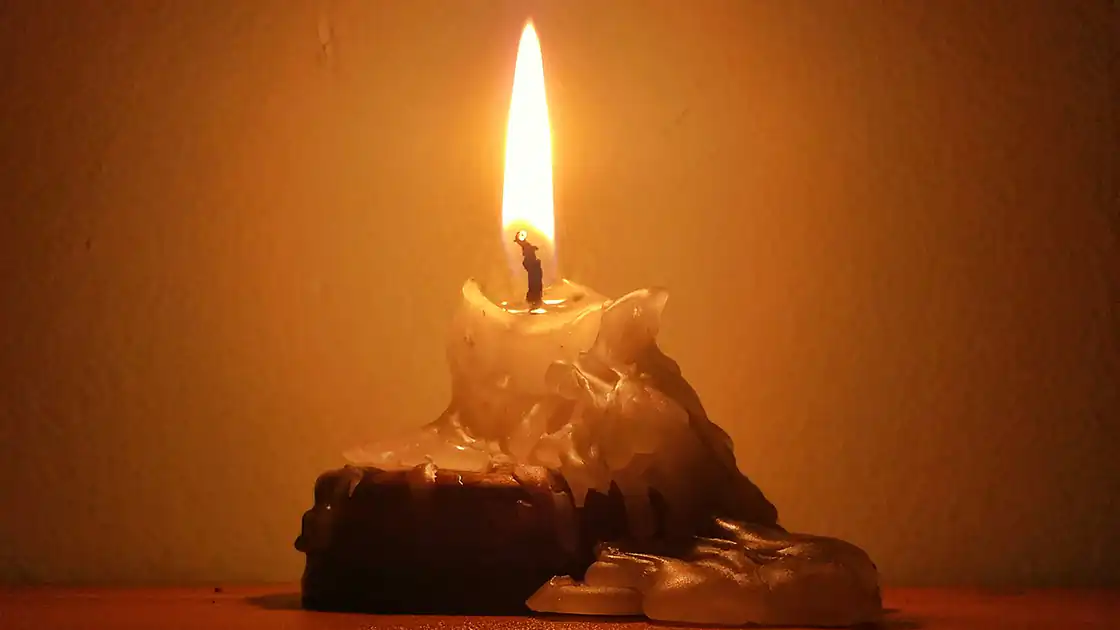 Eine abgebrannte Kerze mit einer großen Flamme ist hier in der Nahaufnahme zu sehen. Das Wachs der Kerze ist bereits herunter geronnen, das Feuer der Flamme erzeugt ein warmes, braun-gelbes Ambietente im Bild.