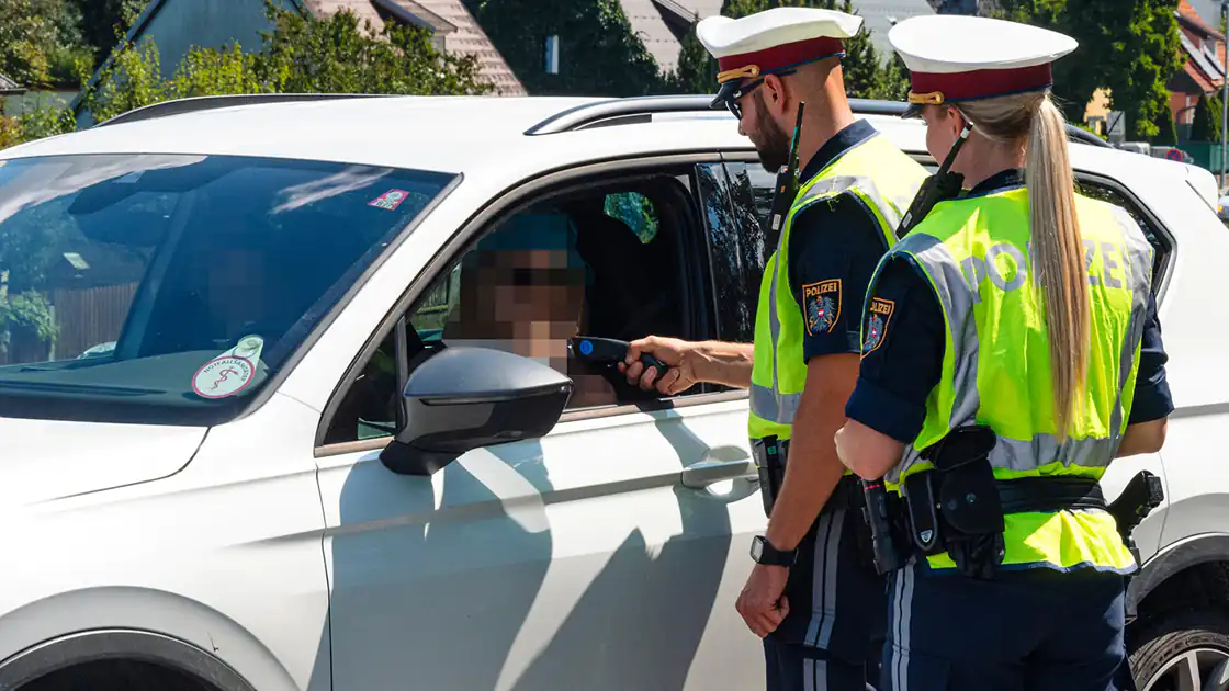 Zwei Polizisten, eine Frau und ein Mann, stehen neben einem weißen Auto. Die Fensterscheibe auf der Fahrerseite des Autos ist geöffnet und der männliche Polizist hält das Alkoholmessgerät durch das seitliche Fenster des Fahrers um einen Test auf Alkohol durchzuführen. Die weibliche Polizistin steht hinter ihm und beobachtet die Szene.