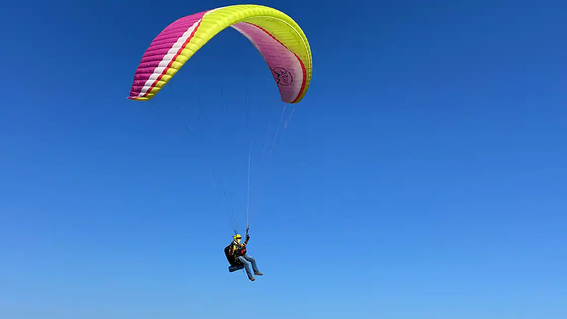 Ein Paragleiter mit Fallschirm ist aus der Entfernung beim Fliegen zu sehen, hinter ihm ist der strahlen blaue wolkenlose Himmel.