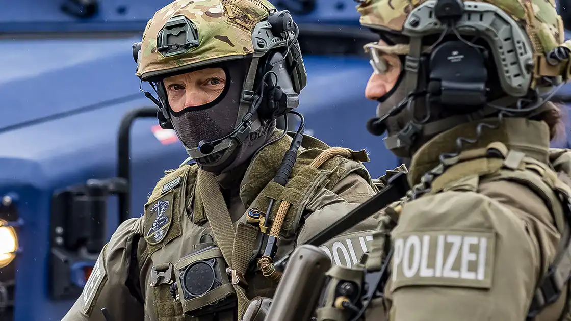In der Detailaufnahme sind zwei Kräfte der Polizei-Spezialgruppe COBRA zu sehen. Die COBRA wird bei Einsätzen mit besonderem Gefahrenpotential beigezogen. Die Spezialkräfte sind schwer bewaffnet mit automatischen Feuerwaffen. Sie tragen Helme in Camouflage-Farben und haben einen Mundschutz und vor dem Mund Mikrophone des Funkgerätes.