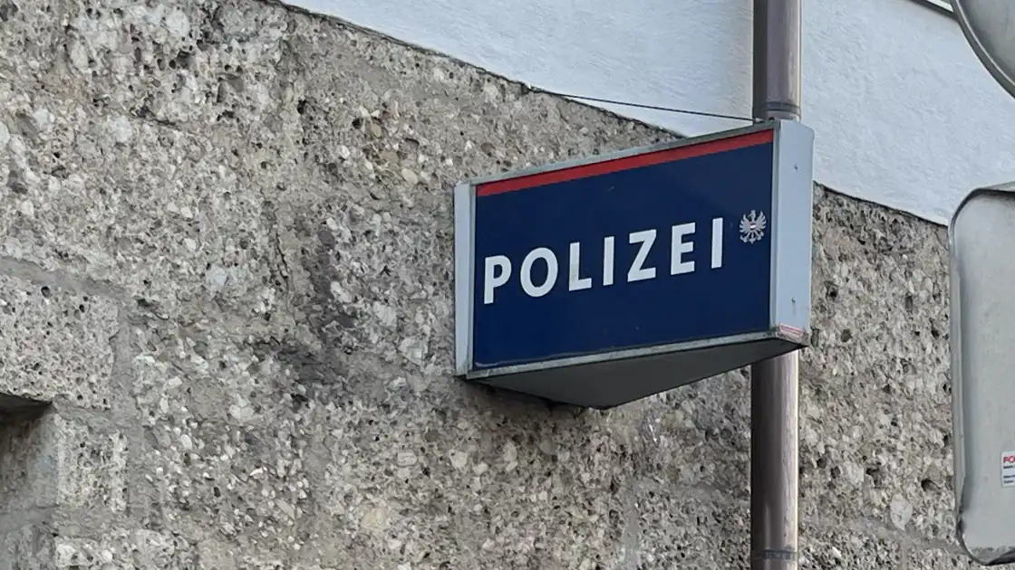 Das Gebäude-Schild einer Polizeistation in Österreich mit einem dreieckigen Leuchtschild, das das Emblem und den Text Polizei trägt. Es ist auf einer Hauswand montiert.