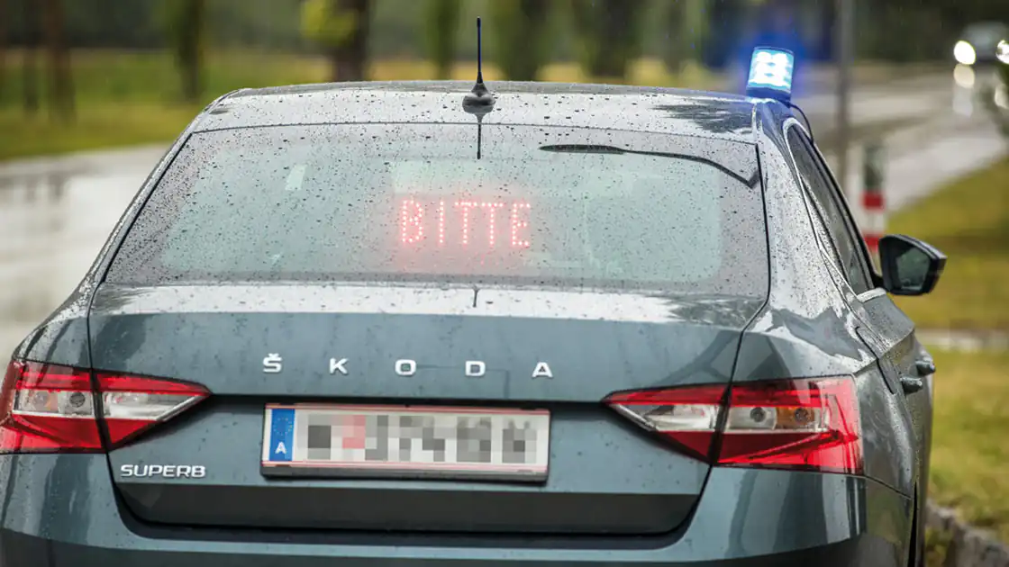 Eine getarnte Zivilstreife der Polizei enttarnt sich. Das privat wirkende Auto hat auf der Beifahrerseite das flexible Blaulicht auf dem Dach stehen, das nur bei bedarf vom Beifahrer hinausgegeben wird. In der Heckscheibe ist eine digitale Anzeige zu sehen, auf der das Wort Bitte steht. Üblicherweise wird der Text dann mit der Aufforderung rechts an den Straßenrand zu fahren, ergänzt.