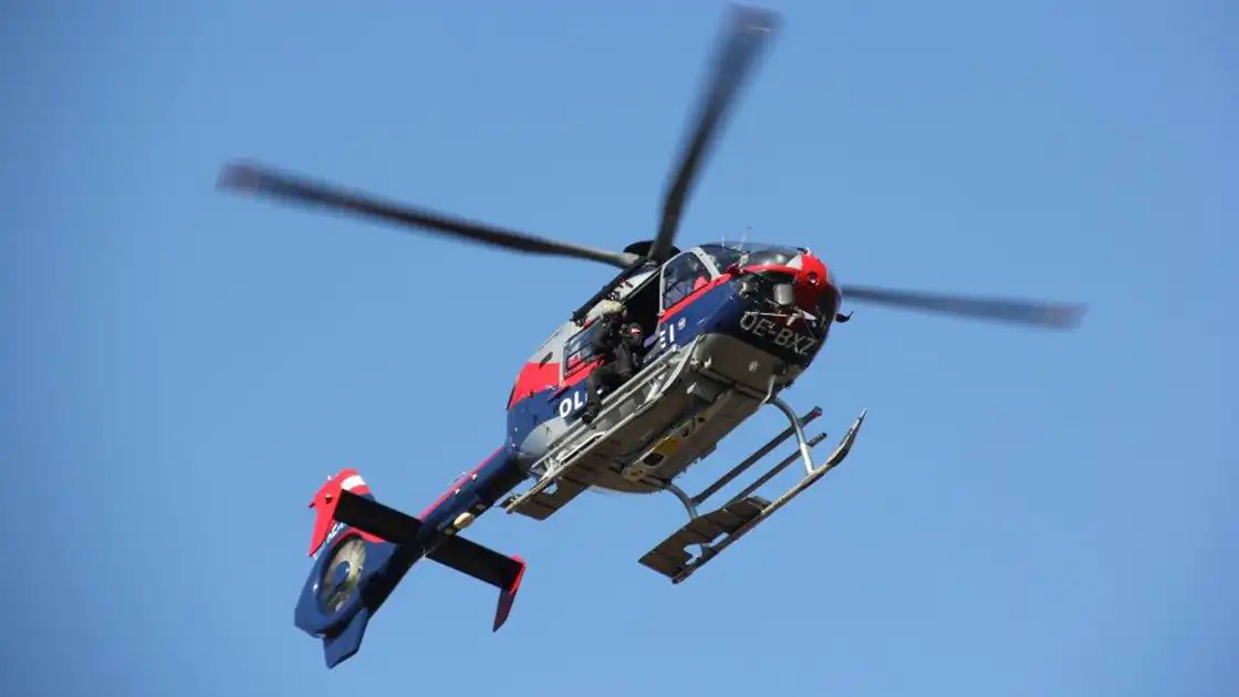 Ein Polizeihubschrauber fliegt in der Luft. Das Bild zeigt den Helikopter der Polizei von unten, im Hintergrund ist lediglich der blaue Himmel zu sehen. Die Tür des Helikopters ist seitlich geöffnet und es ist eine Person an der Seitentür zu sehen, die auf den Kuven des Hubschraubers steht.
