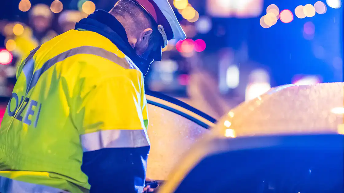 Ein Polizist mit einer gelben, reflektierenden Warnweste bekleidet ist am Abend damit befasst eine Fahrzeugkontrolle durchzuführen. Die Nahaufnahme zeigt den Polizisten neben einem weißen Auto, bei dem die Fahrertür geöffnet ist. Im Hintergrund sind unscharf zahlreiche Lichter aus dem Verkehrsgeschehen, Ampeln und andere Fahrzeuge zu sehen.