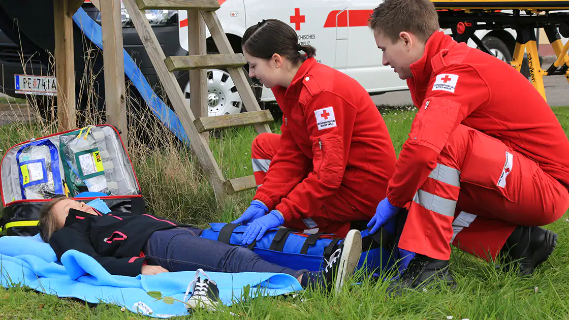 In diesem Symbolphoto ist ein Kind zu sehen, das sich offenbar verletzt hat. Die Umgebung deutet auf einen Spielpatz mit einer Klettermöglichkeit hin, das Kind hat sich am Bein verletzt und wird hier von 2 Sanitätsmitarbeitern geschient und für den Abtransport vorbereitet.