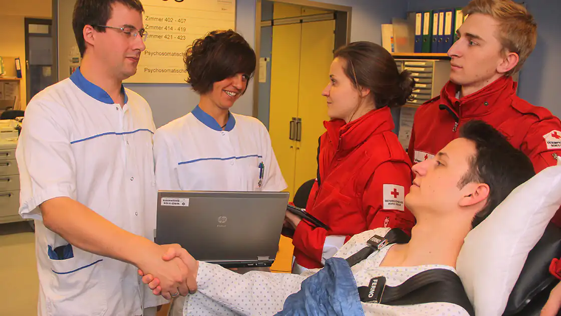 Die Rettung / Rotes Kreuz übergibt einen Patienten im Krankenhaus an die behandelnden Ärztinnen und Ärzte. Ab diesem Zeitpunkt kümmert sich das Krankenhaus um die Patienten. Im Bild ist ein Arzt und eine Krankenschwester zu sehen, die einen Patienten begrüßen, der von zwei Sanitätsleuten ins Krankenhaus gebracht wurde, die auch am Bild sind.