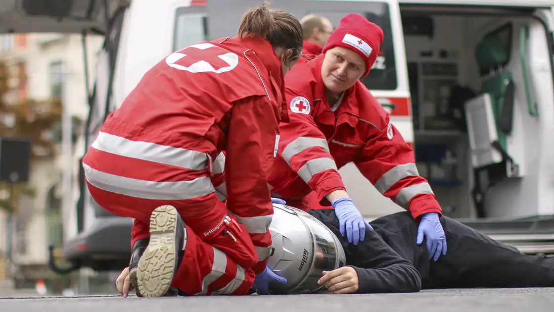 Zwei Sanitäter vom Roten Kreuz die sich um einen Motorradfahrer mit Helm kümmern, der auf dem Boden liegt. Der Motorradfahrer, der verletzt am Boden liegt, hat noch seinen Helm auf dem Kopf. Die Sanitäter sind dabei den Mann zu behandeln.