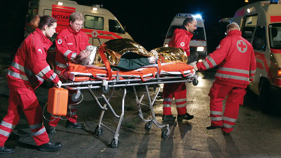Im Bild zu sehen ein Rettungseinsatz mit einem schwer verletzten Patienten. Vier Sanitäter schieben eine Bahre mit einem Patienten in dieser Nacht-Szene zu den Rettungsautos. IM Hintergrund sind drei Rettungsautos erkennbar, der Patient ist in eine goldene Warmhaltefolie eingewickelt.