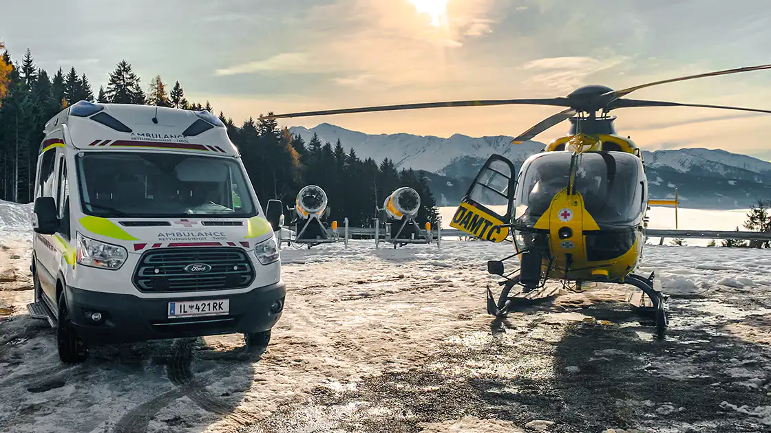 Ein Rettungswagen und ein Hubschrauber sind in winterlicher Landschaft bei einem Skigebiet im Einsatz. Im Hintergrund zu sehen sind auch Schneekanonen.