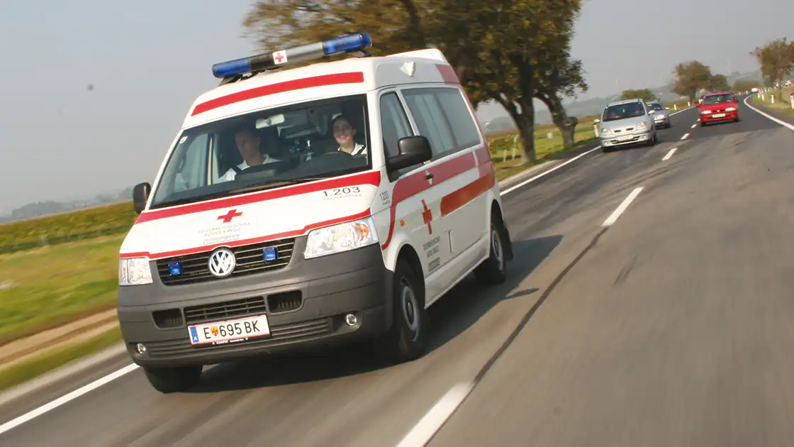 Unterwegs auf einer Landstraße mit zwei Spuren ist ein Rettungsauto im Vordergrund. Dahinter sieht man einige normale PKWs die alle in die gleiche Richtung wie das Rettungsauto fahren, das Fahrzeug der Rettung kommt auf die Kamera zu.