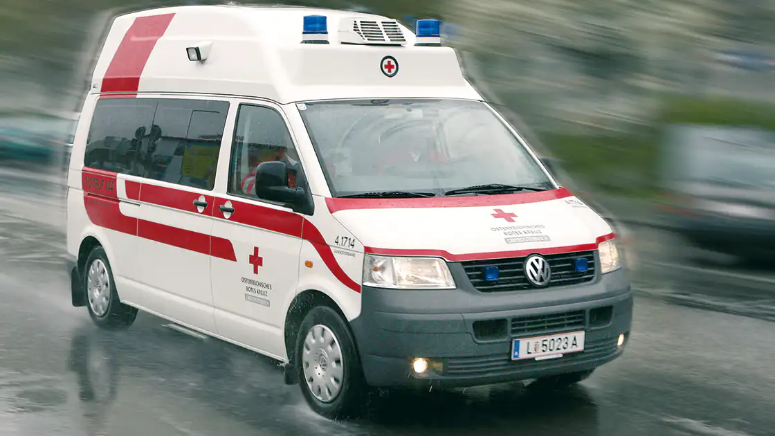 Ein Rettungsauto bei Regen auf nasser Fahrbahn ist unterwegs. Das Rettungsauto ist vollständig im Bild zu sehen.