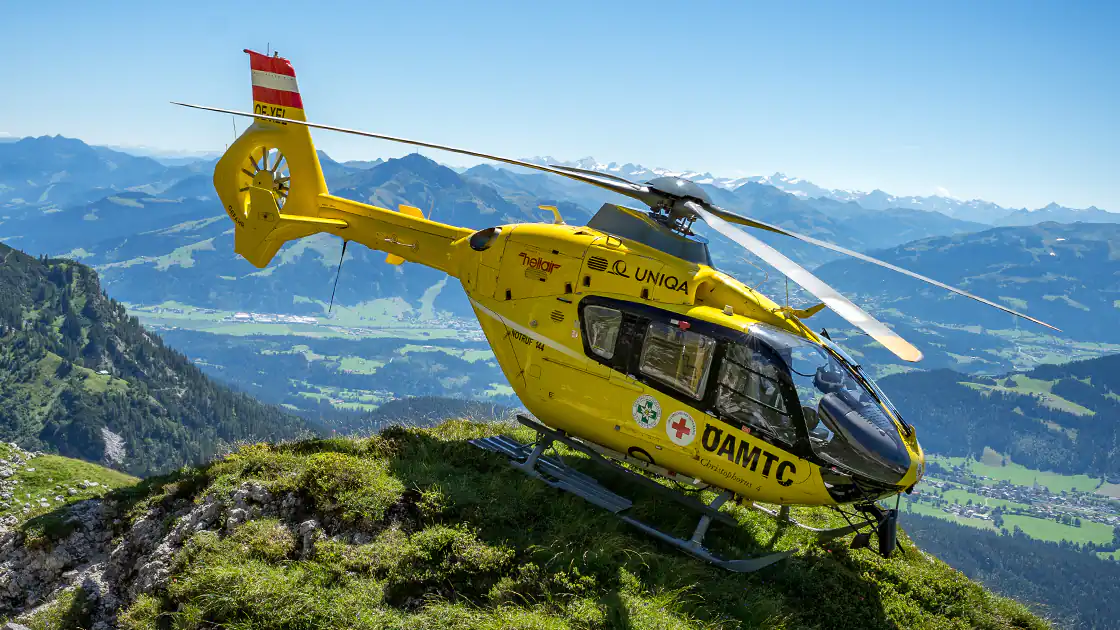 Ein Rettungshubschrauber, der im Gebirge auf einer Bergkuppe geparkt ist, ist im Bild zu sehen. Der Hubschrauber steht schräg auf dem Felsen, im Hintergrund ist ein bewohntes Tal und weitere Berge zu sehen.