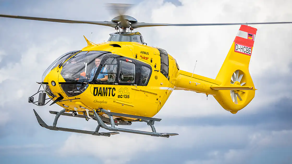 Ein Rettungshubschrauber des österreichischen ÖAMTC ist hier im Flugeinsatz zu sehen. Der Helikopter ist gelb und trägt die rot-weiss-rote Markierung auf der Heckflosse. Im Hintergrund sieht man dicht bewölktes Wetter.