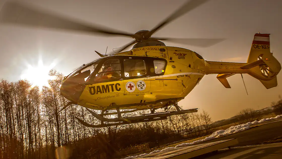 Im Bild ist ein Rettungshubschrauber zu sehen, der vor der Kamera bei untergehender Sonne vorbeifliegt. Der Rettungshubschrauber gehört zur Flotte des ÖAMTC in Österreich.