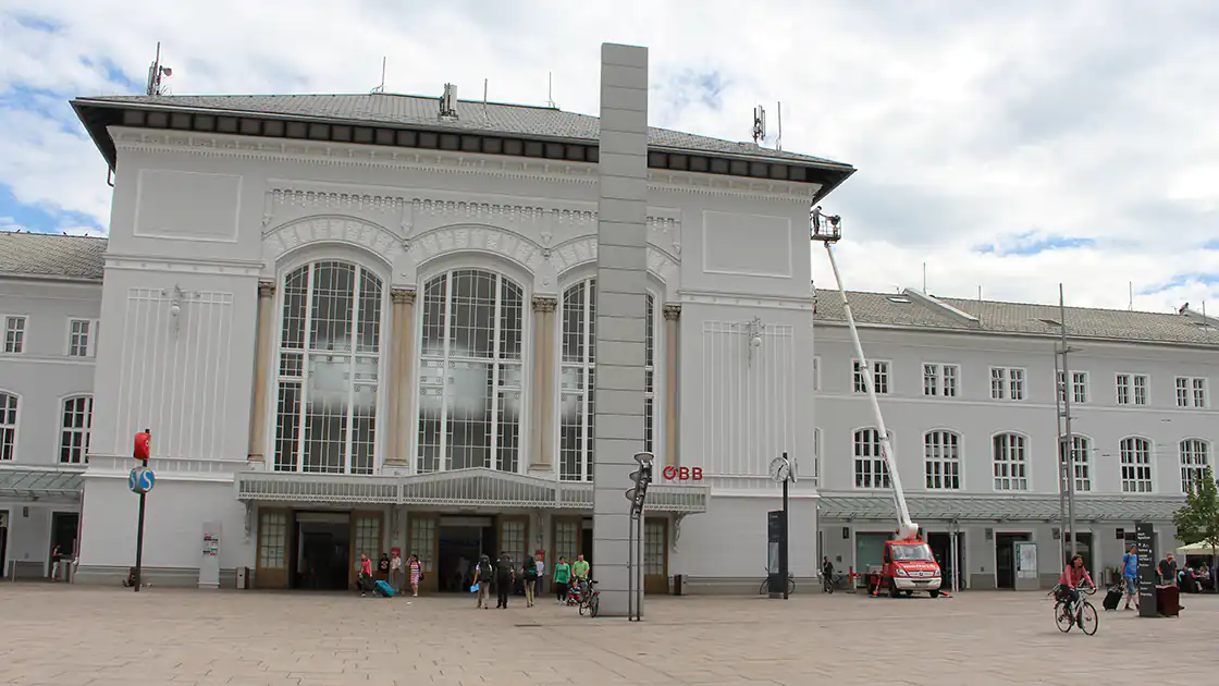 Das Gebäude des Hauptbahnhof Salzburg betrachtet vom Südtirolerplatz aus. Der Bahnhof ist ein großes, weißes Gebäude mit einem Haupttrakt, der höher ist als der Rest des Bahnhofes. Im Bild unten zu sehen ist der Haupteingang zum Salzburg Hauptbahnhof.