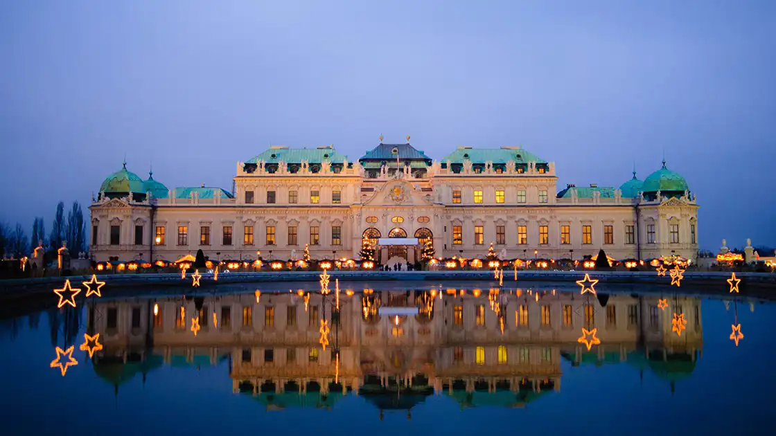 An atmospheric evening view of Schönbrunn Palace in Vienna. The image shows a large expanse of water in front of the palace, reflecting the palace façade as well as lights and torches. The photo was likely taken during the Christmas season, as illuminated star-shaped figures are also visible.
