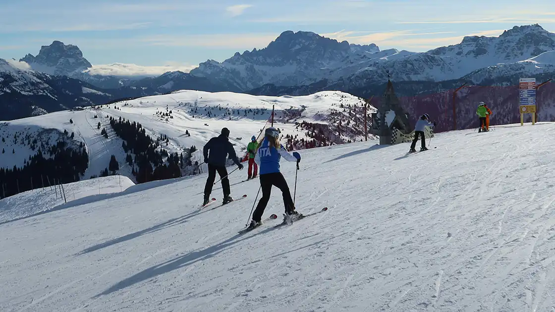 Skifahrer auf einer Skipiste in den Bergen. Auf einem Plateau mit viel Schnee sind mehrere Skifahrer unterwegs, im Hintergrund sind Gebirgszüge zu sehen.