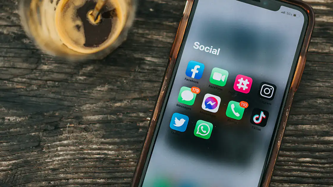 Soziale Netzwerke, zumeist auf Smartphones wie hier im Bild genutzt, aber auch auf klassischen Computern oder Tablet-Geräten. Neben einem Glas mit einem dunklen Getränk liegt ein Smartphone mit mehreren Icons von sozialen Netzwerken auf dem Bildschirm. 
