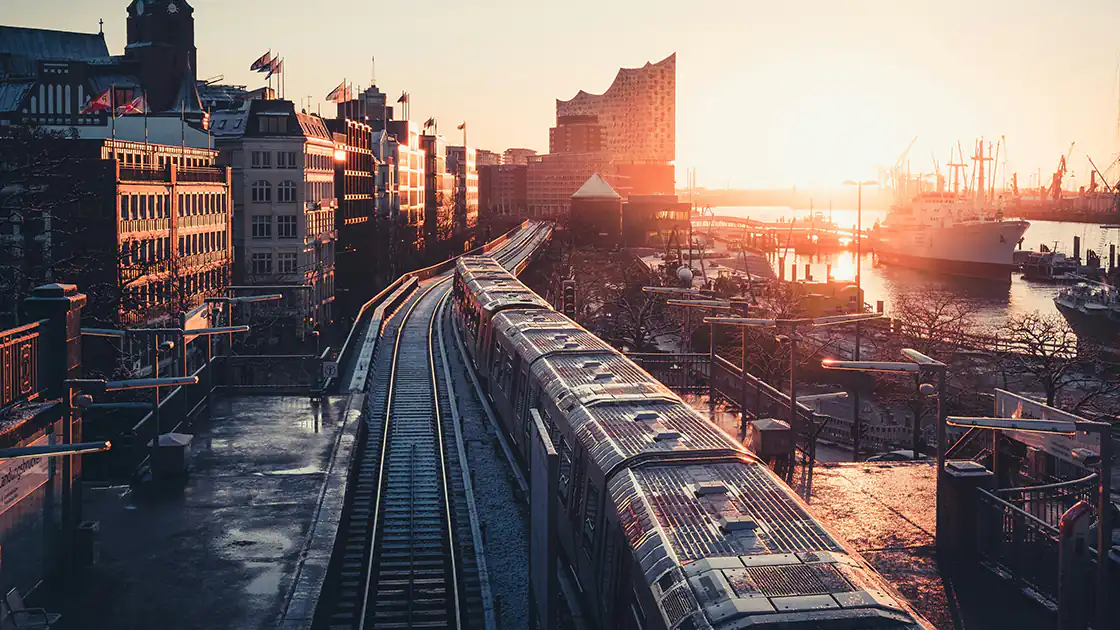 In einer Großstadt ist eine Bahntrasse gelegt. Man sieht Gleise und einen langen Zug, der entlang der Gleise geführt wird. Die Gleise führen neben einigen Häusern vorbei, die Stadt ist typisch europäischen Stils, die Häuser sind nur einige Stockwerke hoch. Im Hintergrund ist der Sonnenuntergang zu sehen.