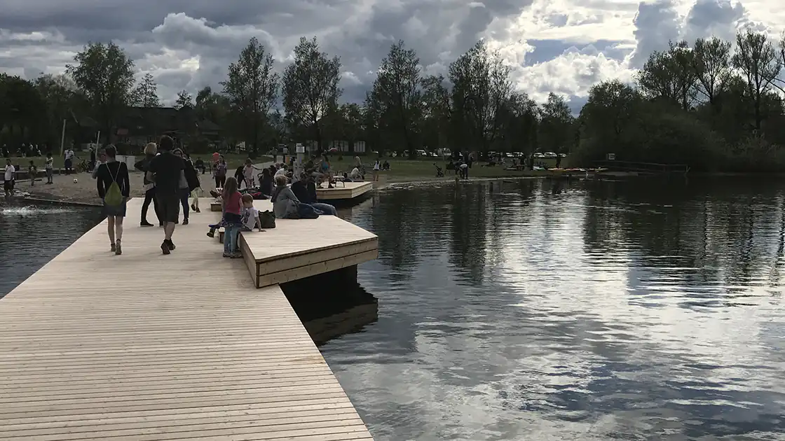 Im Bild zu sehen ist ein Holzsteg, der sich an einem Badeplatz / Strandbad an einem See befindet. Auf dem Steg sind einige Menschen zu sehen, das Bild wurde vom Steg in Richtung Seeufer aufgenommen. (Das konkrete Bild zeigt das Strandbad in Seekirchen am Wallersee.)