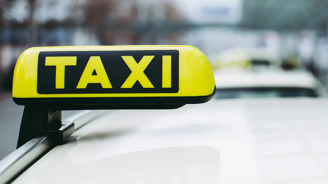 Nahaufnahme eines Taxi-Schildes auf dem Dach eines Taxi-Autos. Das gelbe Leuchtschild hat eine schwarze Fläche und wiederum Gelb das Wort Taxi aufgedruckt. Es ist eine Tagesaufnahme, das Auto hat eine helle beige Farbe.