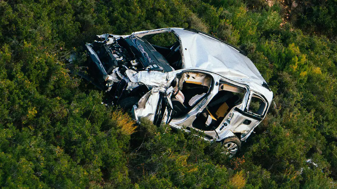 Ein völlig zerstörtes Fahrzeug nach einem Unfall liegt schräg auf einem Waldhang. Das Auto ist an der Front vollständig zerstört, die Fenster sind herausgebrochen, die Autotüren sind herausgebrochen und das Dach des Autos ist eingedrückt. Das Auto dürfte sich beim Abstürzen auf dem Hang offenbar mehrmals überschlagen haben.