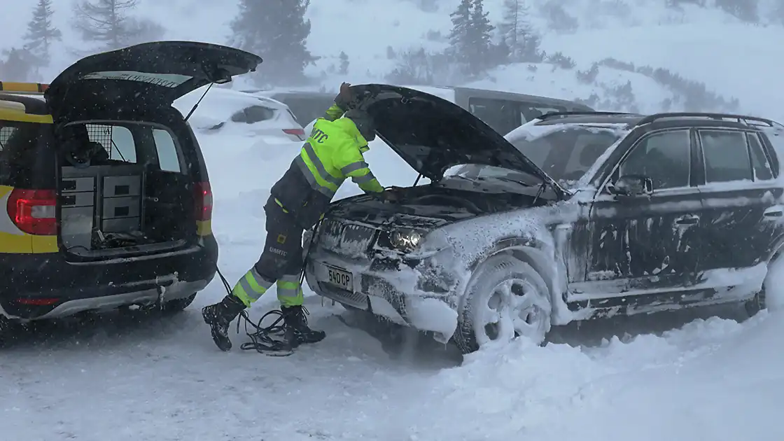 Pannenhilfe vor Ort im Winter. Ein zugeschneites und teilweise gefrorenes Auto wird von einer Person vom Pannendienst wieder in Gang gesetzt. Die Person steht vor der geöffenten Motorhaube des Fahrzeuges. Das Pannenhilfe-Fahrzeug steht daneben und hat den Kofferraum geöffnet. Bei dieser Pannenhilfe wird gerade Starthilfe für die Batterie des Pannenfahrzeuges geleistet.