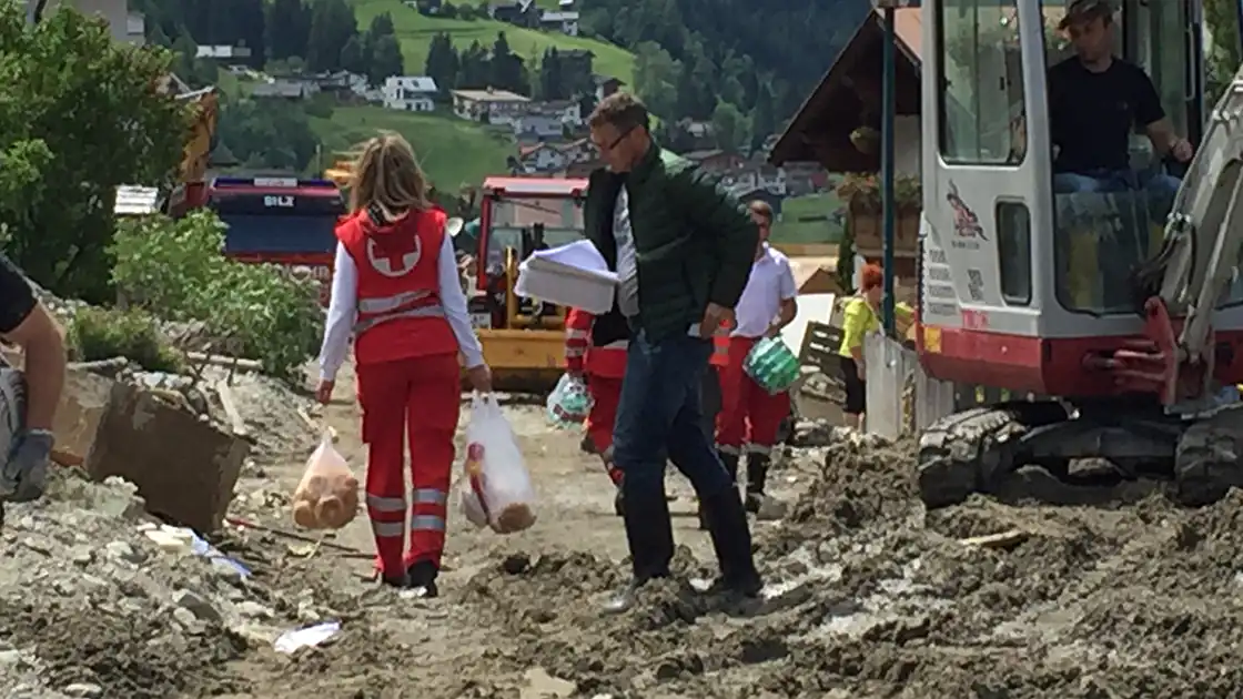 Im Bild zu sehen sind Rettungskräfte bei Aufräumarbeiten bei einer Naturkatastrophe / Überschwemmung / Murenabgang in einer ländlichen Region. Daneben ist auch ein kleiner Bagger bei den Aufräumarbeiten der mit Schotter und Geröll zugeschütteten Straße zu sehen.
