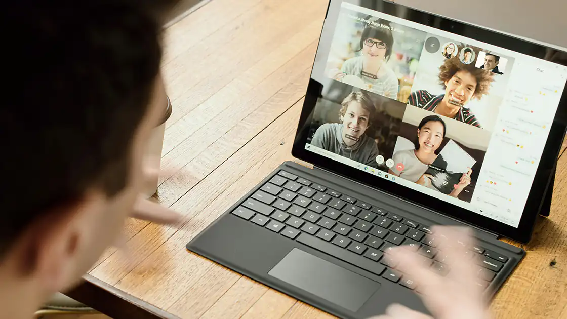 Auf dem Bildschirm dieses Mannes, der vor der Kamera sitzt, ist die Szene einer Videokonferenz mit vier Personen auf einem Laptop zu sehen. Der Laptop steht auf einem braunen Holztisch.