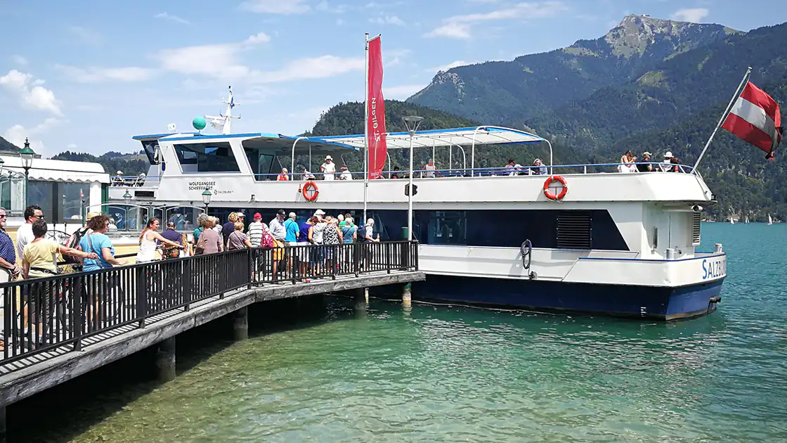 Schifffahrtsstation am Wolfgangsee: Eine Personenfähre hat am Ufer / Hafen angelegt und Menschen auf dem Steg gehen zum Schiff. Das Schiff fährt am Wolfgangsee im Salzkammergut. Hinten am Schiff ist eine österreichische Flagge zu sehen. Im Hintergrund sieht man die Berge am Wolfgangsee.