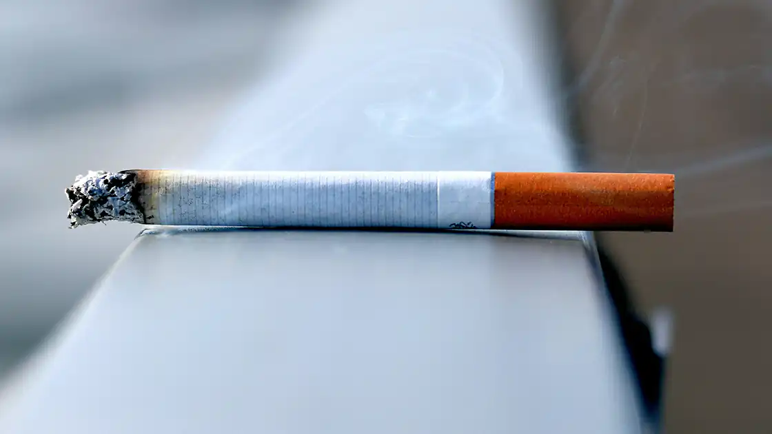 Auf einem Metallgeländer liegt eine brennende Zigarette, die vorne bereits Asche hat. Der entstehende Rauch ist im Bild kaum erkennbar.