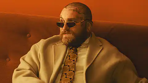 Teddy Swims ist der Künstlername des US-amerikanischen Künstlers und Singer-Songwriters, der mit bürgerlichem Namen Jaten Collin Dimsdale heißt. Auf dem Bild ist der Mann mit Glatze und zahlreichen Tätowierungen im Gesicht und am Kopf auf einer gepolsterten Couch sitzend zu sehen. Er trägt ein Hemd, eine Krawatten mit Blumen und eine dunkle Sonnenbrille sowie einen Trenchcoat. Der Künstler hat auch einen Vollbart.