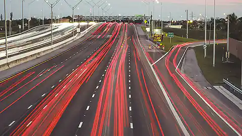 Ein Symbolbild für Autobahnverkehr ist dieses Bild. Man sieht eine mehrspurige Autobahn mit einer Ausfahrt. Die Autos sind nicht zu erkennen, es handelt sich um eine Aufnahme mit langer Belichtungszeit, daher sind nur die roten Lichter als lang gezogene Streifen sichtbar. Auf der Gegenfahrbahn sieht man dementsprechend viele weiße Lichtstreifen von den Autos, die der Kamera entgegen gefahren sind.