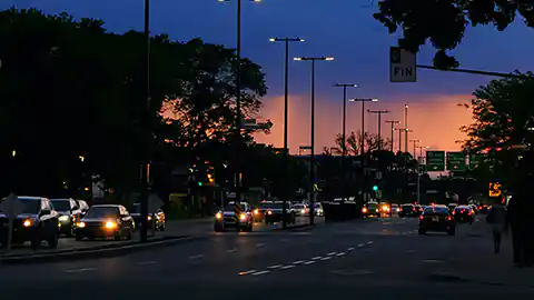 Eine mittelmäßig stark befahrene Straße ist in der frühen Nacht zu sehen. Im Hintergrund ist noch sichtbar, dass die Sonne vor kurzer Zeit untergegangen ist, die Autos stehen in der Schlange und fahren in zwei Richtungen. Die Straßenlaternen sind eingeschaltet.