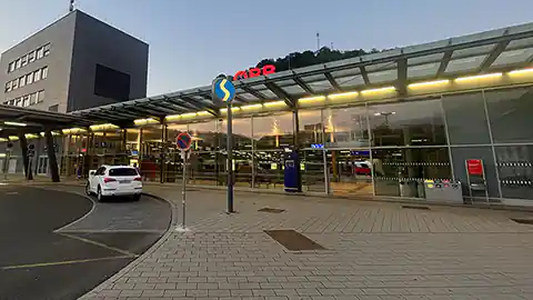 Im Bild zu sehen das Hauptgebäude vom Bahnhof Bruck an der Mur in der Steiermark. Der Bahnhof ist ein wichtiger Umsteigebahnhof für Verbindungen im Dreieck zwischen Wien, Graz und der Weststeiermark sowie folgend Salzburg und Klagenfurt. Im Vordergrund steht ein Masten mit einem S-Bahn-Hinwies, der Bahnhof wirkt modern, hat eine große Glasfront und viel Beleuchtung. Das Gebäude ist aber nur ein Stockwerk hoch. Auf der linken Bildseite ist die Zufahrt für Taxis und Autos zu sehen.