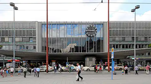 Der Bahnhof München Hauptbahnhof mit dem Haupteingang davor. Vor dem Gebäude des Bahnhofes stehen Taxis und es sind Menschen auf der Straße. Die Mitte des Gebäudes hat eine hohe Fensterfront mit einer großen analogen Uhr darauf. Unter der Fensterfront befindet isch eine große Überdachung. Links und rechts vom Haupteingang befinden sich Geschäfte am Hauptbahnhof München.