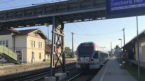 Der Bahnhof Seekirchen am Wallersee mit einem einfahrenden Zug und einem Hinweisschild über die nächste Abfahrt nach Salzburg. Über dem Zug ist ein Fußgängerübergang zu sehen, der es möglich macht auf die andere Seite des Bahnhofes zu gelangen.