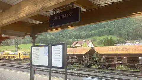Dieses Bild ist ein Symbolbild für den Bahnhof Unzmarkt in der Steiermark, man sieht einen Bahnsteig mit dem Schild für Unzmarkt und dahinter mehrere Gleise und ein Gleis, auf dem sich ein Güterzug mit Waggons für Schüttgut befindet.